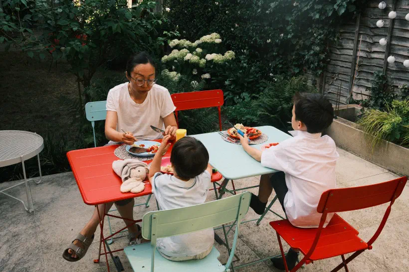 Three people sit at two tables in a garden. There is food on the table in front of them. A fourth place is laid out but no one sits there.