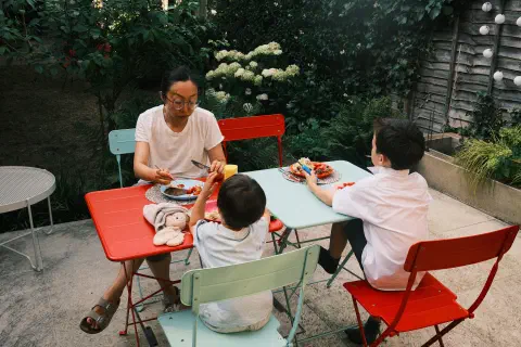 Three people sit at two tables in a garden. There is food on the table in front of them. A fourth place is laid out but no one sits there.