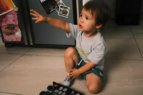 A young boy kneels on the floor reaching up to someone off camera. Behind him is a fridge, in front of him on the floor is a pack of frozen sesame buns.