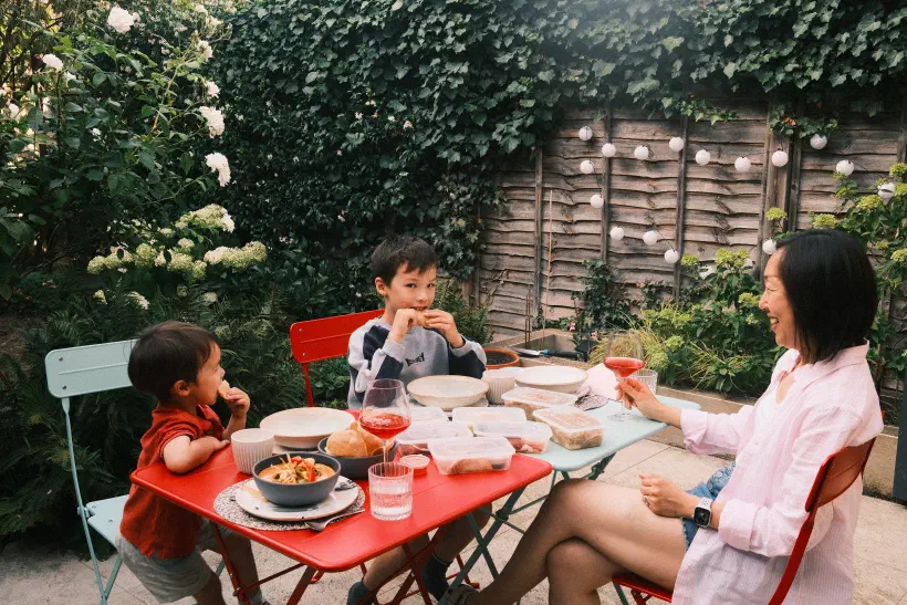 A family sits around an outdoor table in a garden. On the table is food and a glass of wine. A lady holds a glass of wine smiling at two young boys.