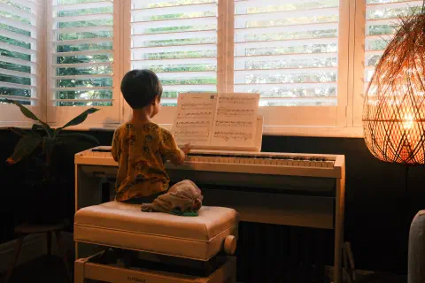 A young boy sits at the piano in front of brightly lit windows. On the stool to his right, a soft rabbit sits next to him. In front of him some music, his hands on the keyboard.