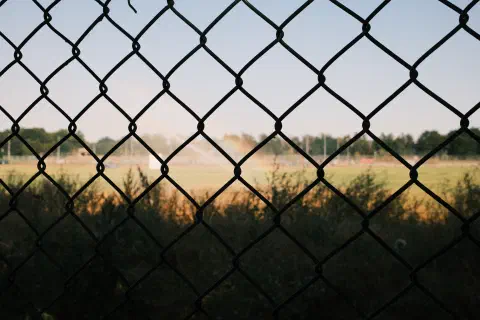 A photo taken through some wire fence. Behind the fence, in the shade, some wild grass. In the distance, brightly lit playing fields. A subtle rainbow appears in the spray of a sprinkler.