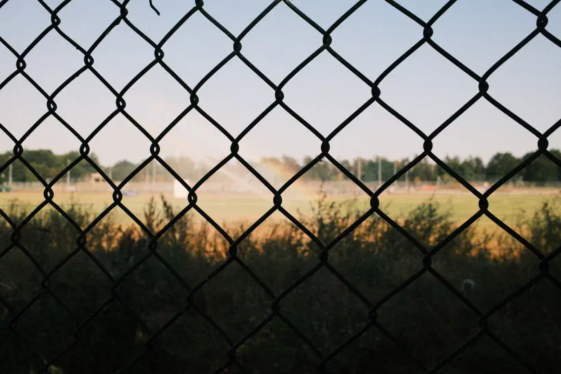 A photo taken through some wire fence. Behind the fence, in the shade, some wild grass. In the distance, brightly lit playing fields. A subtle rainbow appears in the spray of a sprinkler.
