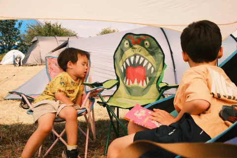 Two boys sit on camping chairs, the younger of the two is fast asleep. In between them a chair that looks like a dinosaur.