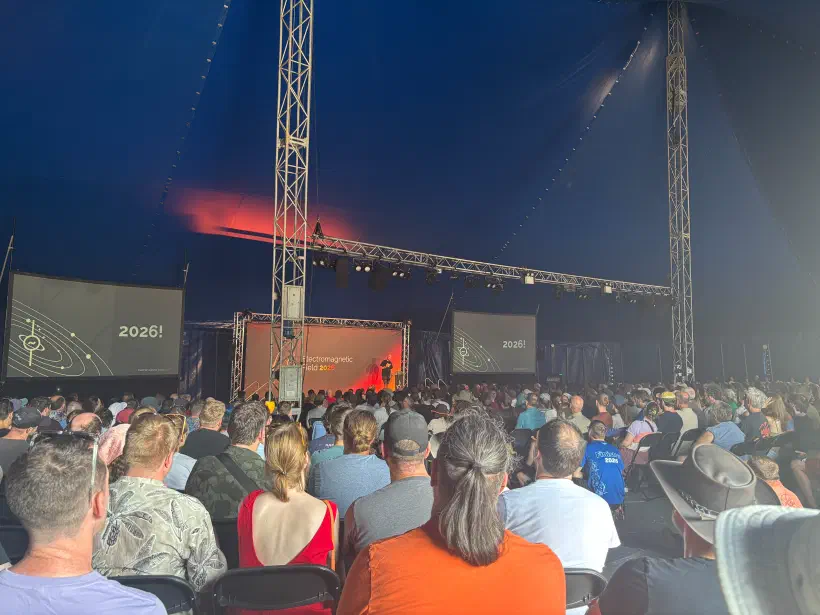 A photo of a marquee tent filled with a crowd of people looking towards a stage. Either side of the stage two large screens. A person stands on stage. Behind them a background, says Electromagnetic Field