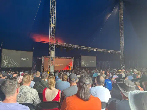 A photo of a marquee tent filled with a crowd of people looking towards a stage. Either side of the stage two large screens. A person stands on stage. Behind them a background, says Electromagnetic Field