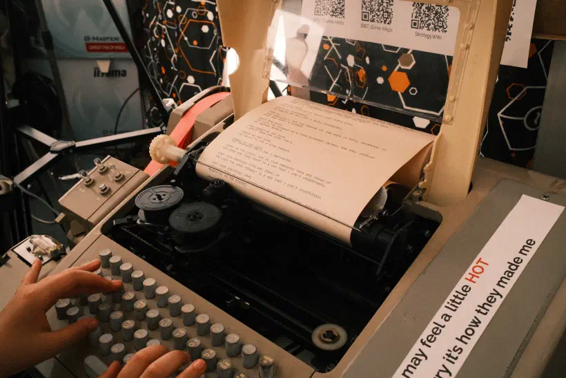 A photo of a teletype machine two hands type on the keyboard in front of the machine several QR codes provide links to an explanation of the game on the piece of paper that appears from the machine instructions a text-based adventure game