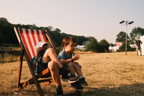 Two boys sit on deck chairs in an otherwise empty looking field. The grass is dry and yellow