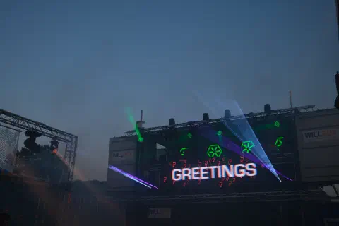 A nighttime photo looking up at a DJ booth above a dance floor. Lights shine towards the photographer. A screen on the front of the booth has the word greetings in white letters.