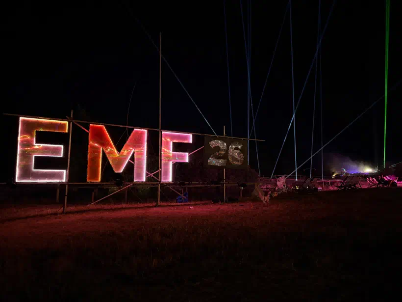 A nighttime photo of the EMF logo. The letters 26 can be seen beside the logo, decorated with little tiny stars. Behind the sign, searchlights and lasers light the night sky