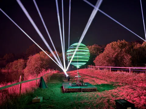A slow-motion photo captures a spinning globe of LEDs. Behind the globe, searchlights light up the sky. The foreground is lit with a pink glow