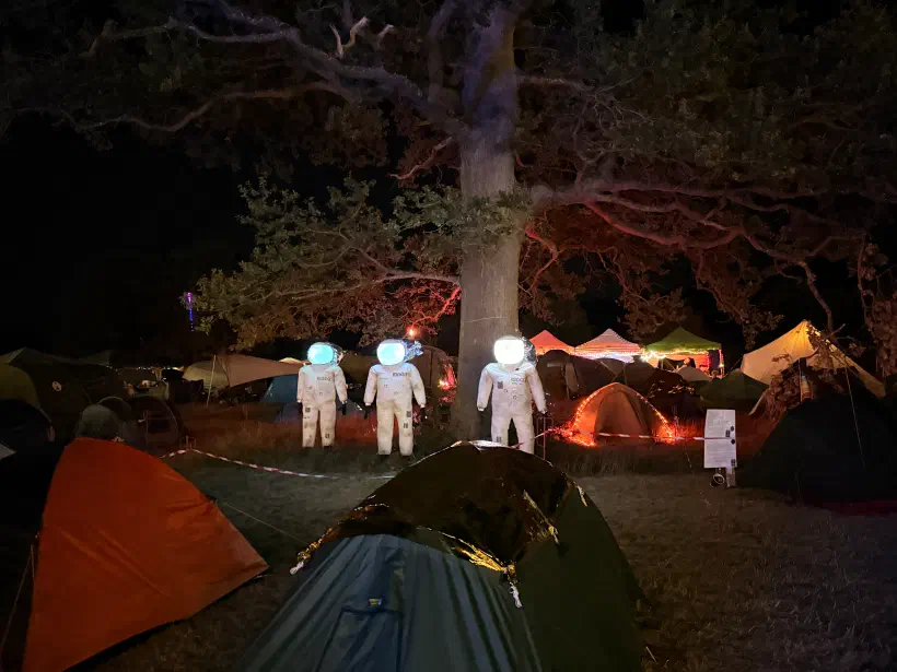 Three figures in spacesuits with brightly lit faceplates stand in a field of tents