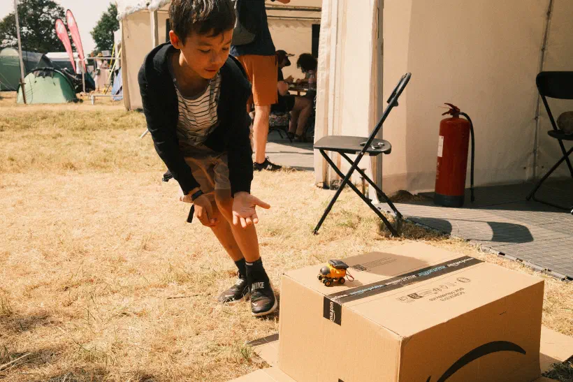 A photo of a boy rushing to catch a small tiny robot that looks like it's about to fall off the edge of an upturned box. The box is on dry grass, outside, a tent.