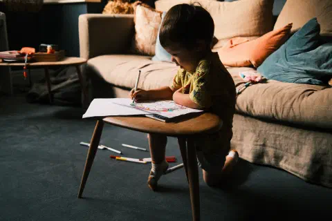 a young boy kneels at a coffee table in front of a sofa, pen in hand, colouring, on the floor behind him, a scattered collection of coloured pens, light from the windows off camera means the majority of the boys figure is silhouted against the bright background