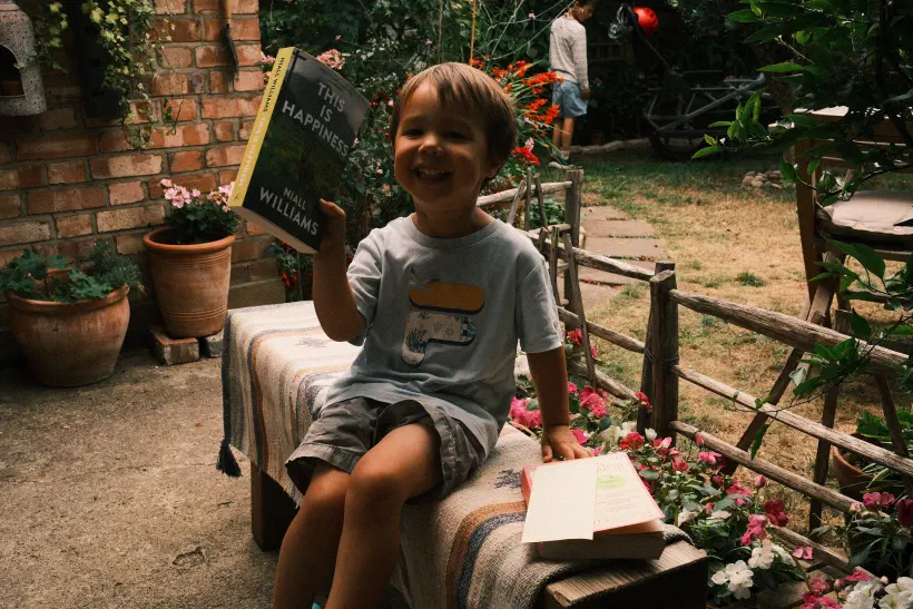 A young boy sits on a garden bench. Behind him, a wooden fence separates the patio area from dry looking grass, in the background another boy stands in the shadows, the boy in the foreground is holding up a book titled, 'this is happiness'.