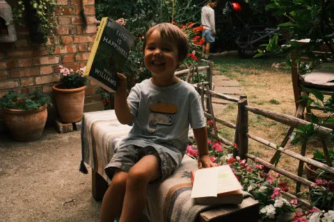 A young boy sits on a garden bench. Behind him, a wooden fence separates the patio area from dry looking grass, in the background another boy stands in the shadows, the boy in the foreground is holding up a book titled, 'this is happiness'.