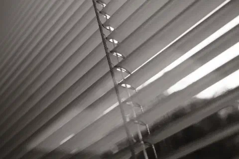a balck and white photo looking out through the slats of a blind to the bright sky outside.
