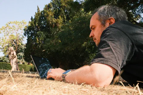 A person lays on dry grass, a laptop in front of him.